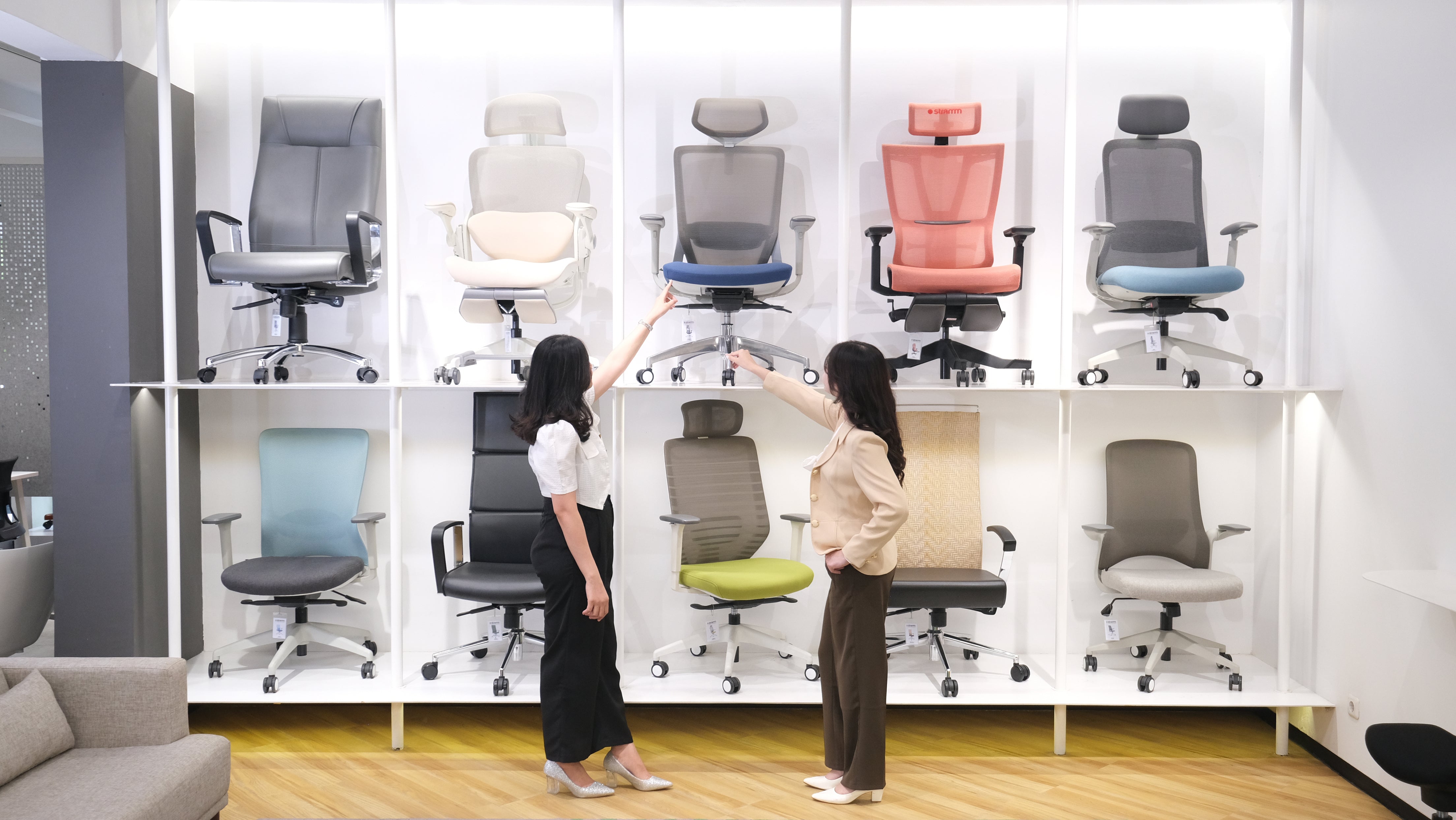 Dua orang sedang melihat dan membandingkan berbagai model kursi kantor ergonomis yang dipajang di showroom furnitur modern. 