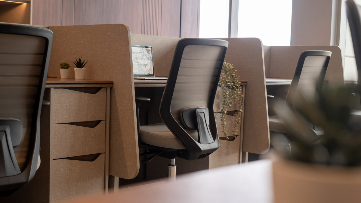 Suasana kantor modern dengan deretan kursi kerja ergonomis berwarna hitam dan meja kerja berpartisi cokelat, dilengkapi laci penyimpanan dan dekorasi tanaman hias.