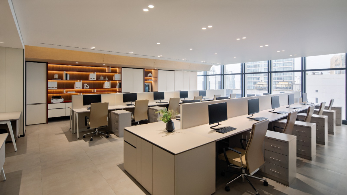 Interior kantor modern dan luas dengan deretan meja kerja putih, monitor komputer, kursi kantor ergonomis, jendela besar, serta rak kayu dengan pencahayaan hangat di bagian belakang.